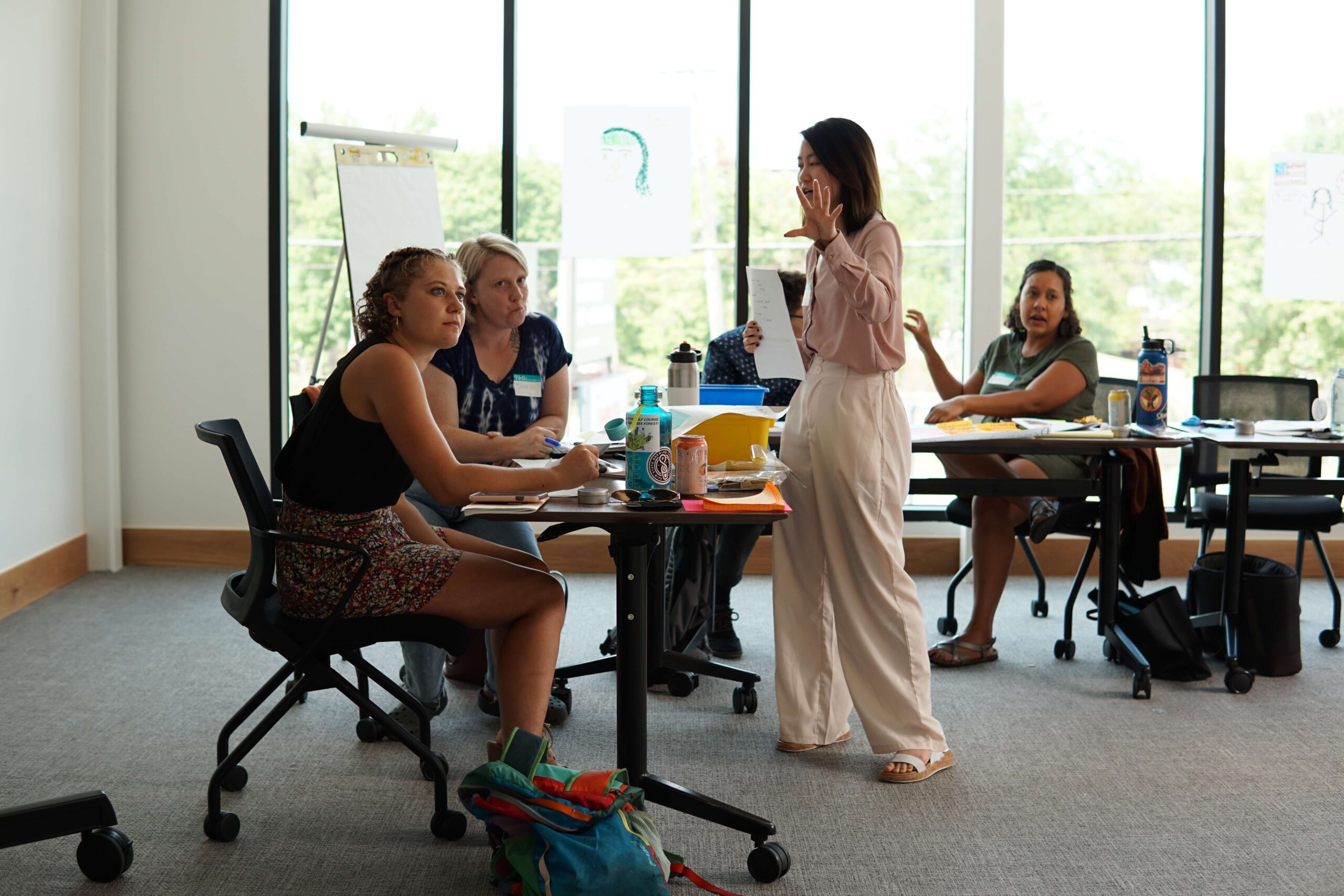 A woman stands and speaks to a group of people seated at tables in a room with large windows.