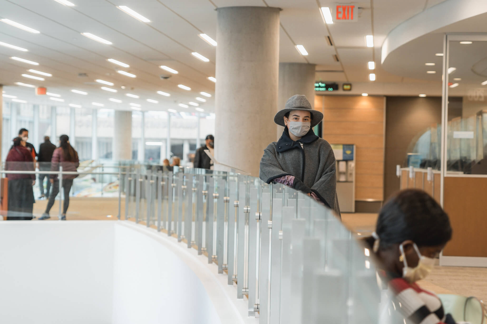 A person wearing a mask stands in a modern, well-lit hallway with large windows.