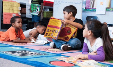 Children sitting on a colorful rug in a classroom, one child reading a book.